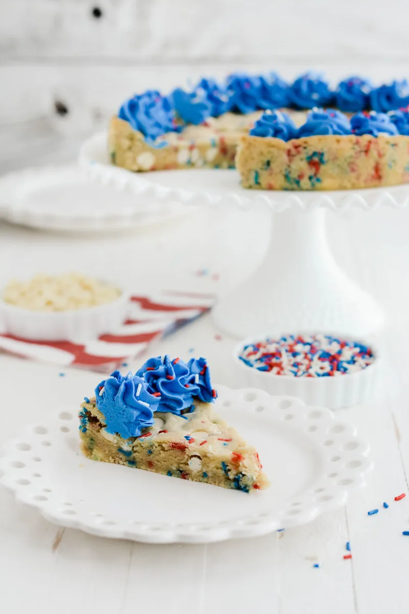 Wide open shot of July 4th Cookie Cake with slice on a plate and remaining cookie cake on a cake pedestal.