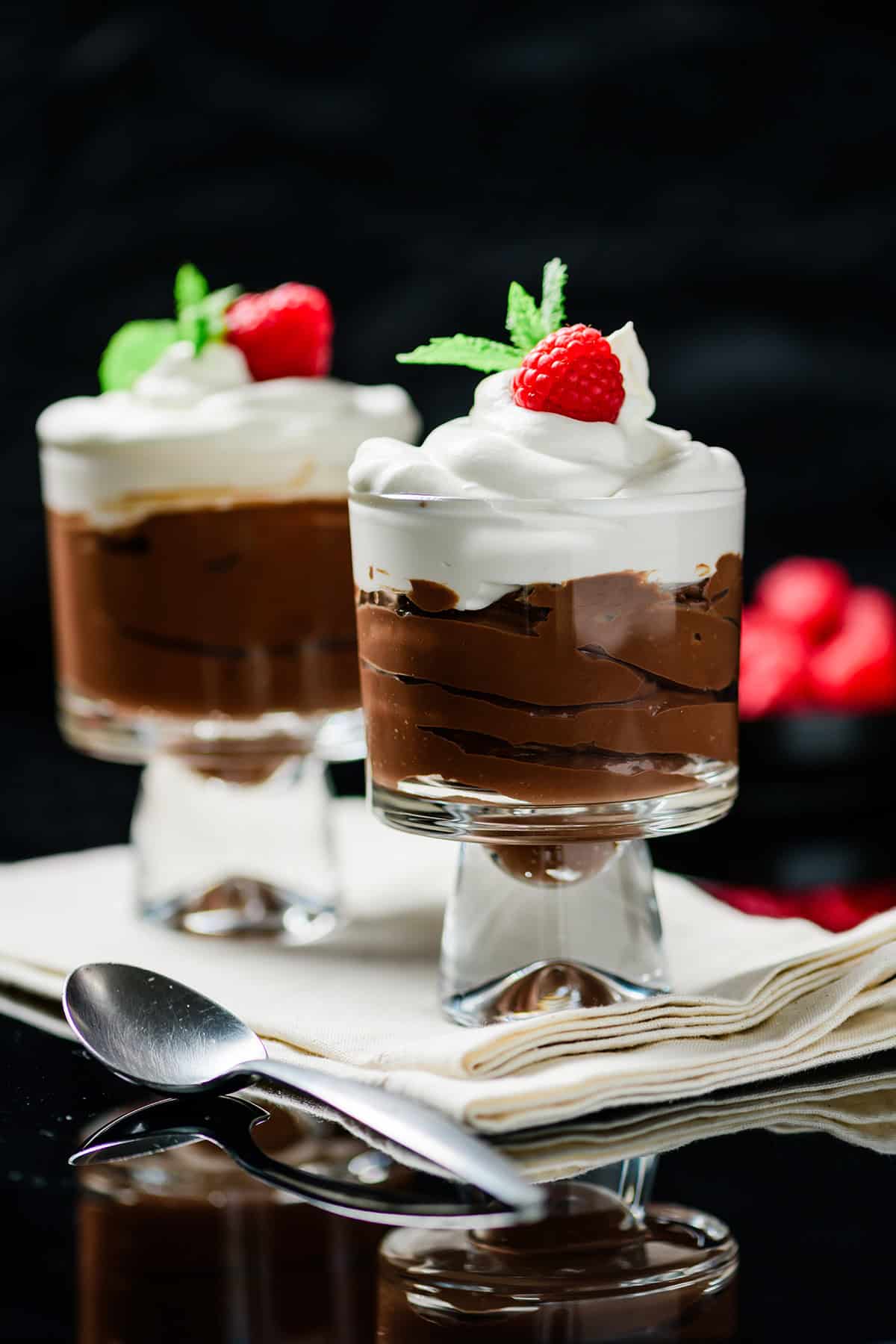 Two parfait glasses filled with Protein Packed Chocolate Mocha Pudding with a spoon and a bowl of raspberries.