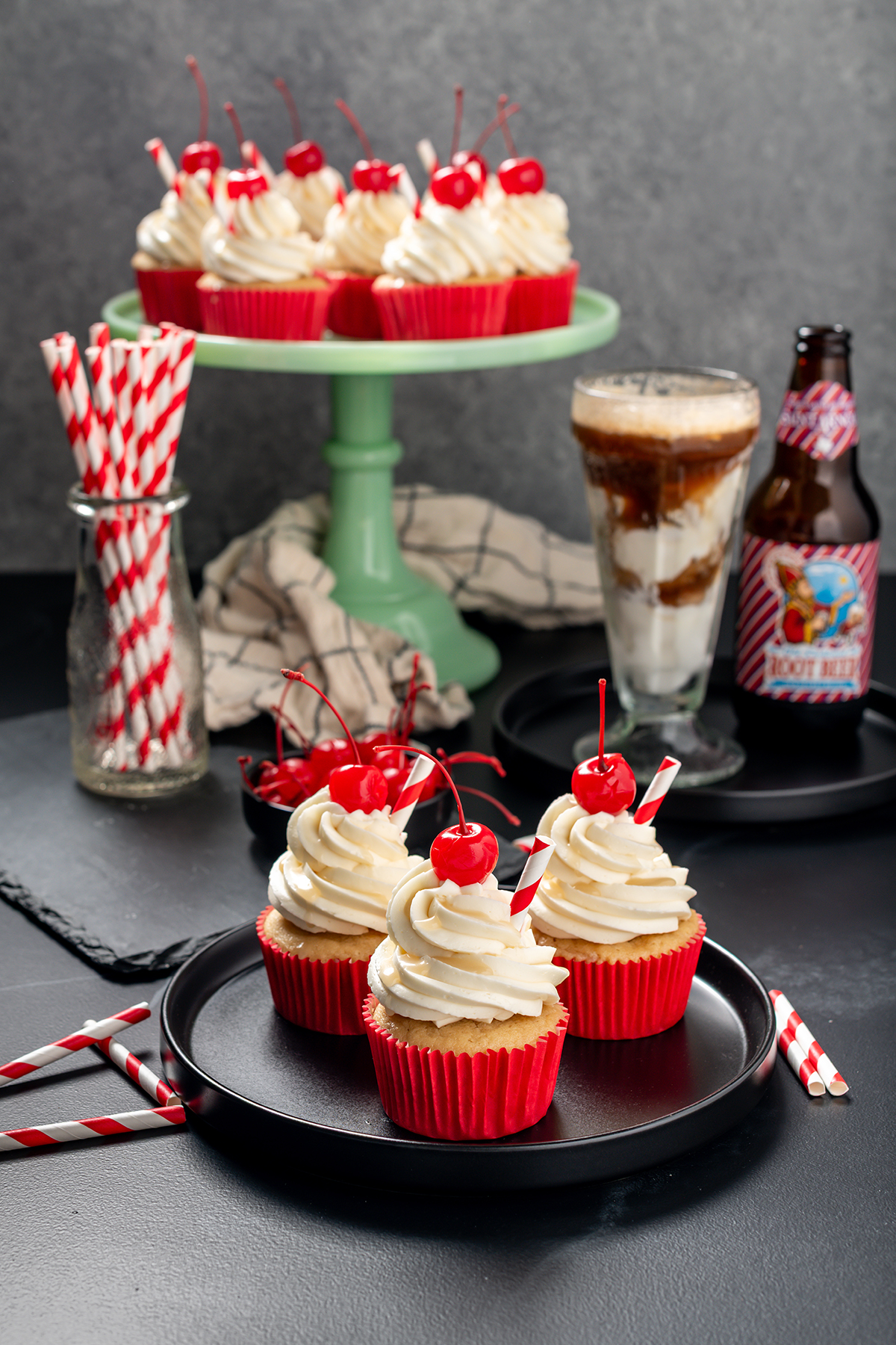 Wide open shot of Root Beer Float Cupcakes on a black plate with cupcakes on a green cake stand in the background.
