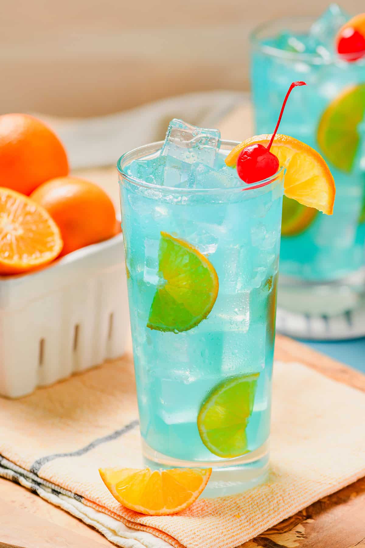 Close up of one Blue Lagoon Cocktail at a slight three quarter angle, with second cocktail in the back with a basket of oranges.