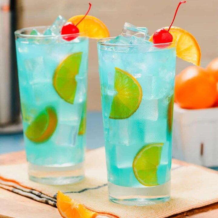 Two Blue Lagoon Cocktails head on shot. with oranges and cocktail shaker in the background.