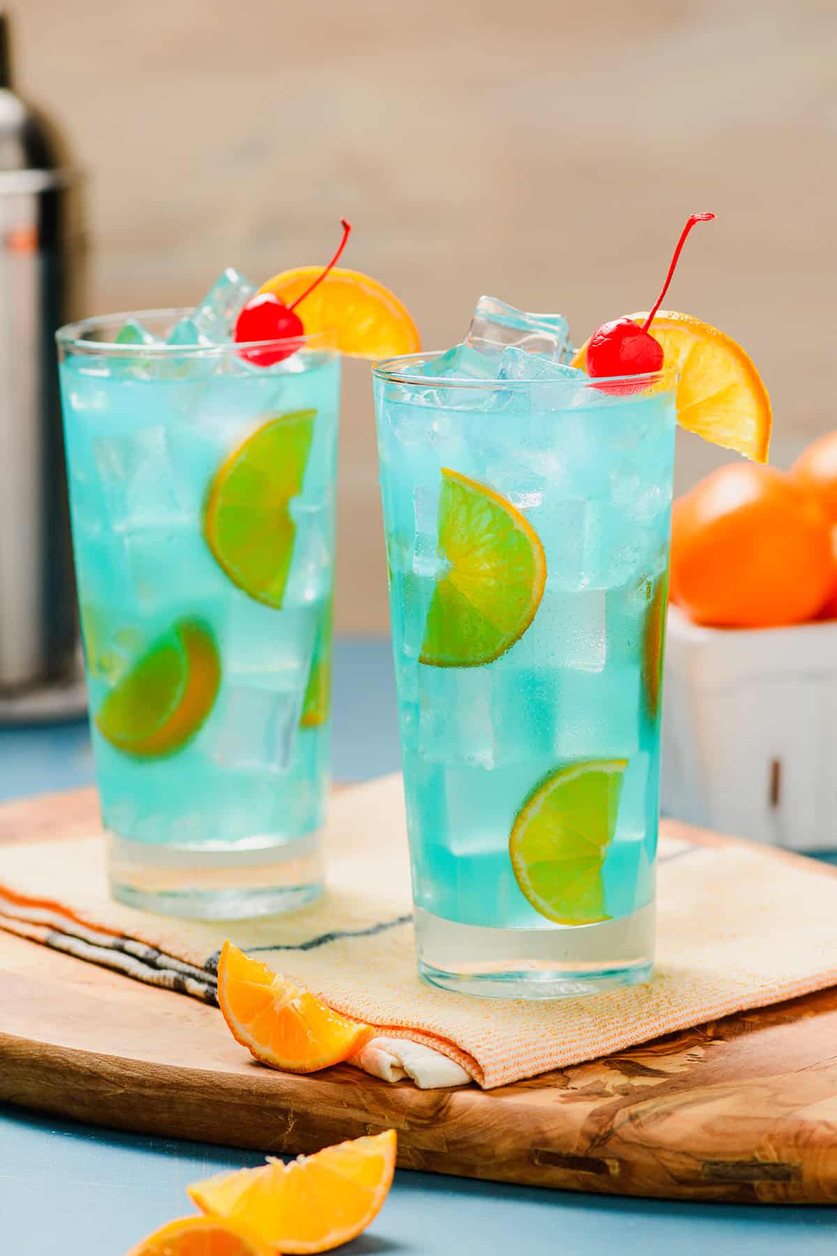 Two Blue Lagoon Cocktails head on shot. with oranges and cocktail shaker in the background.