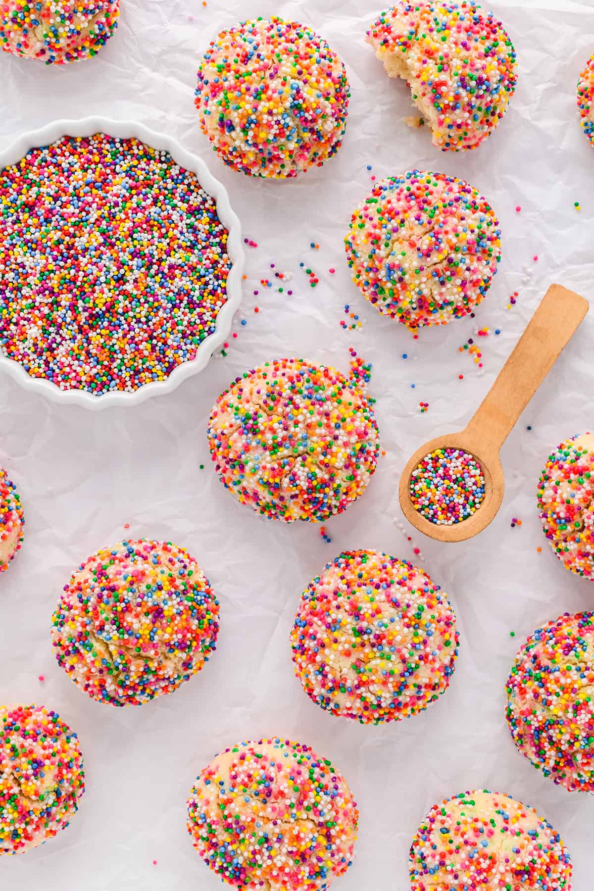 Overhead shot of Mexican Sprinkle Cookies  on top of wrinkled parchment paper with a bowl of sprinkles and a spoon of sprinkles.