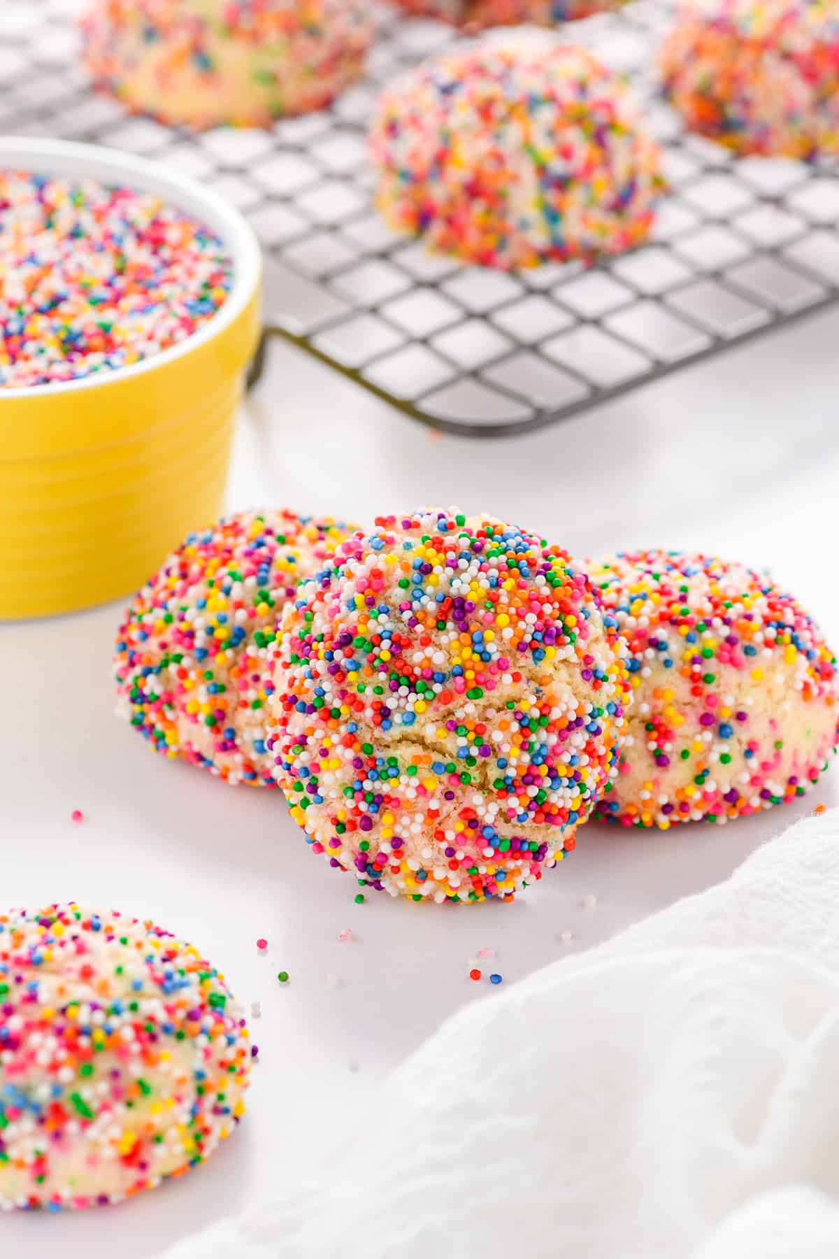 Angled shot of a pile of three Mexican Sprinkle Cookies with a yellow bowl of sprinkles in the background.