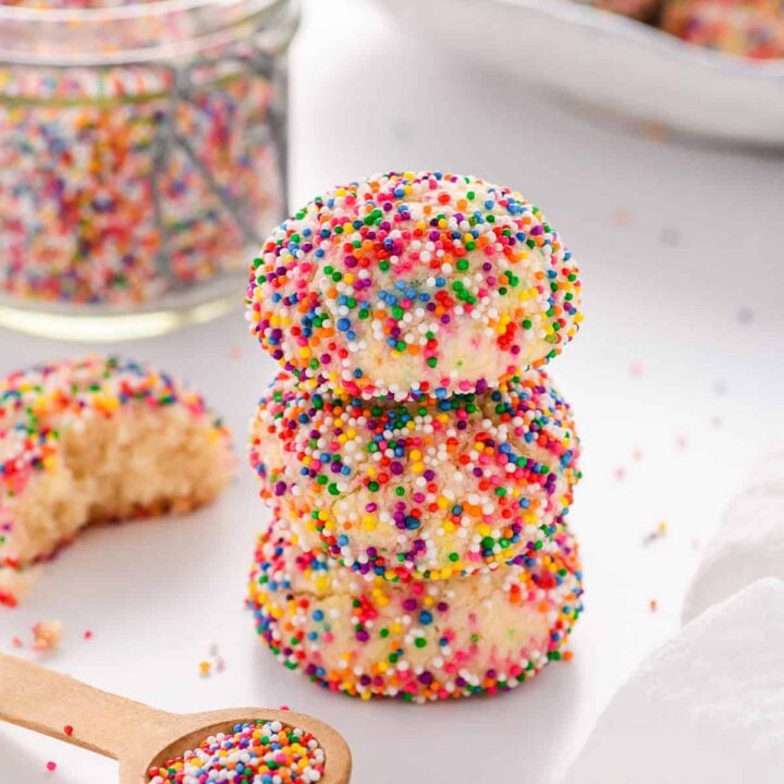 Angled shot of a stack if three Mexican Sprinkle Cookies with a small spoon of sprinkles, jar of sprinkles and a plate of cookies in the background.