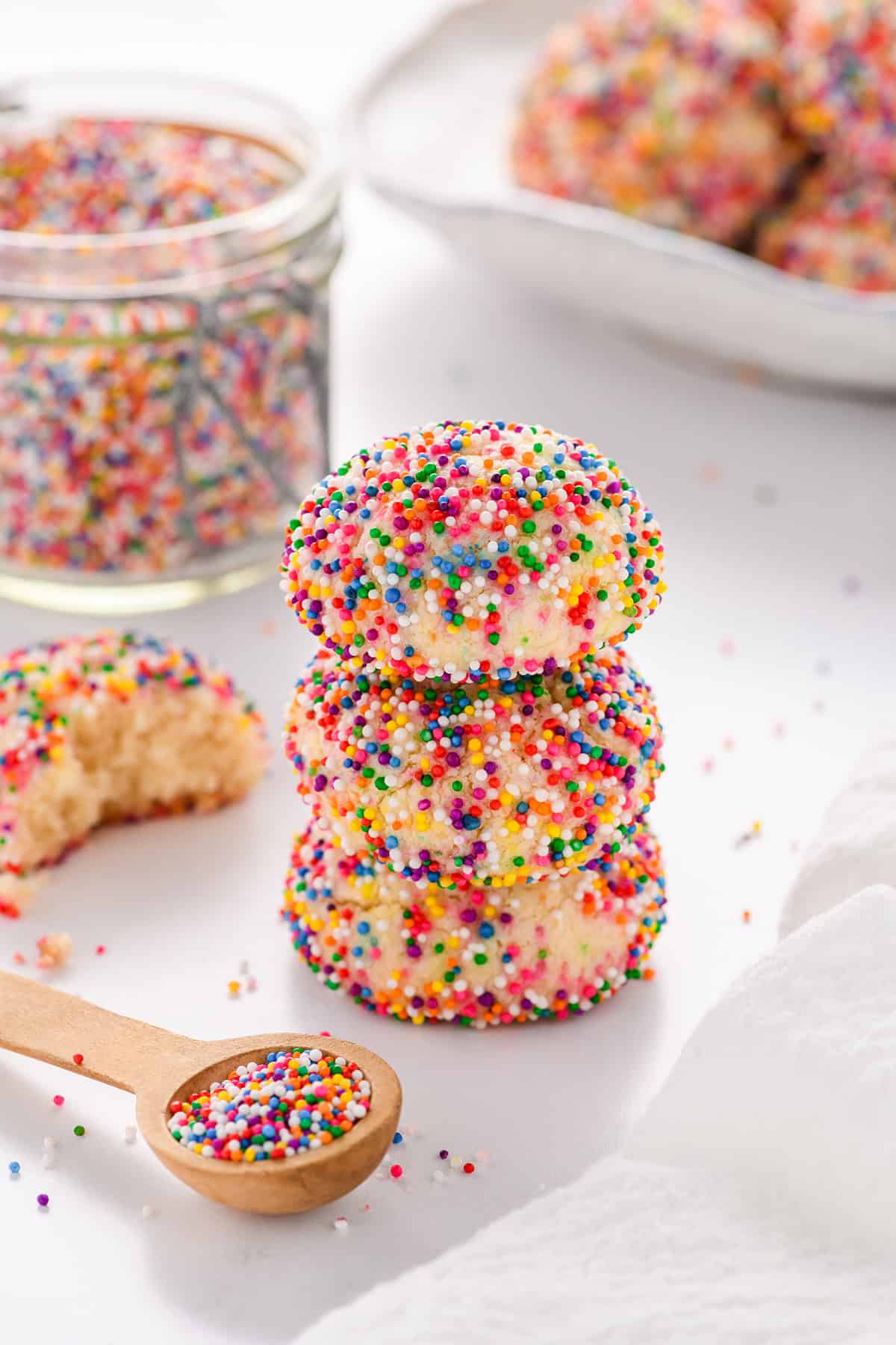 Angled shot of a stack if three Mexican Sprinkle Cookies with a small spoon of sprinkles, jar of sprinkles and a plate of cookies in the background.