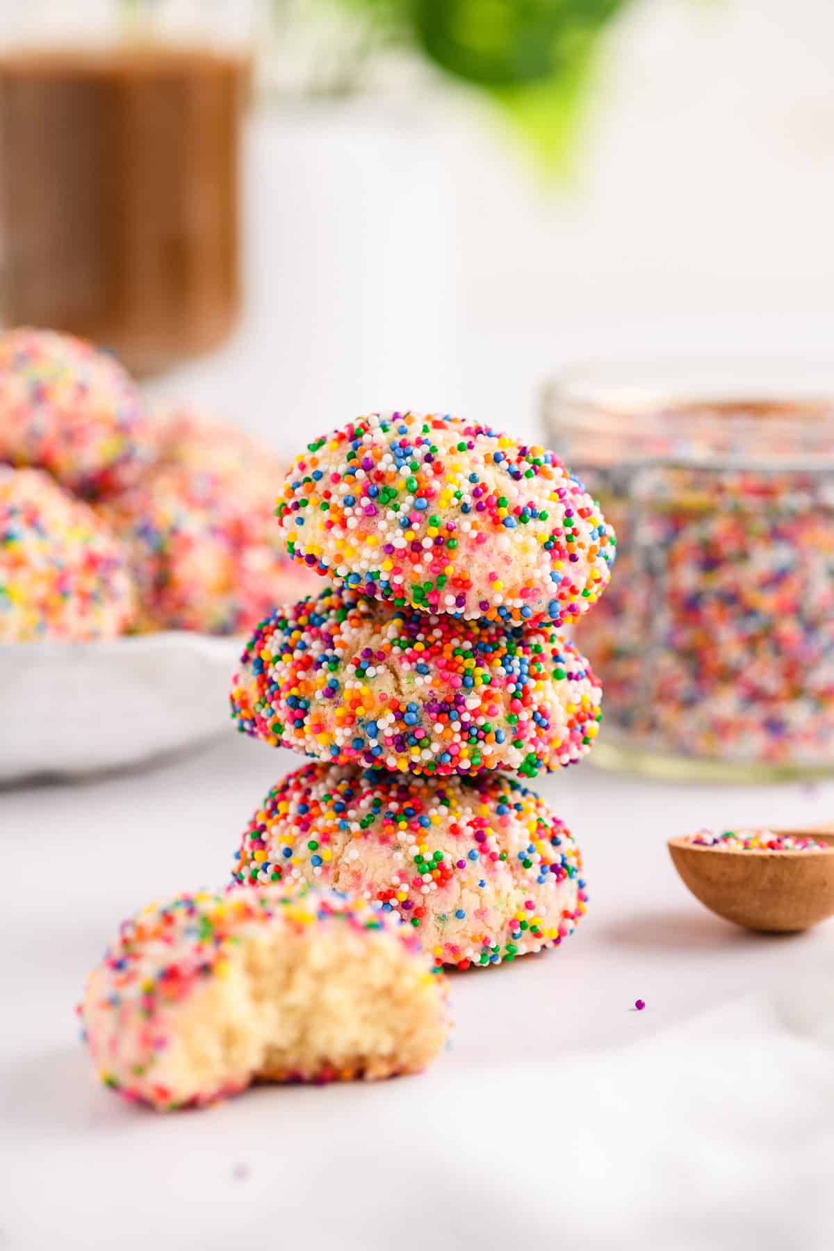 Head-on shot of 3 stacked Mexican Sprinkle Cookies with plate of cookies in the background with coffee and jar of sprinkles.