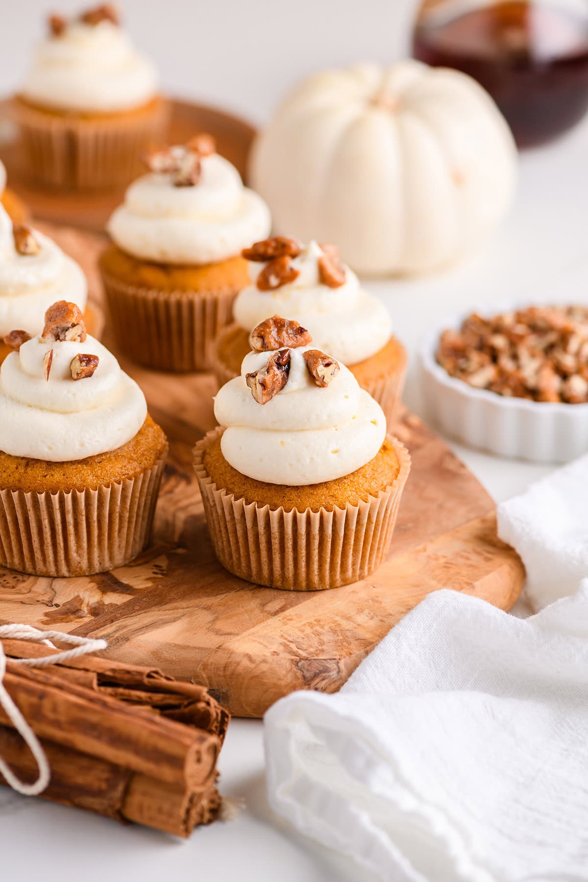 Pumpkin Cupcakes with Maple Cream Cheese Frosting on wood serving board with mini pumpkin and a bowl of chopped candied pecans in the background.