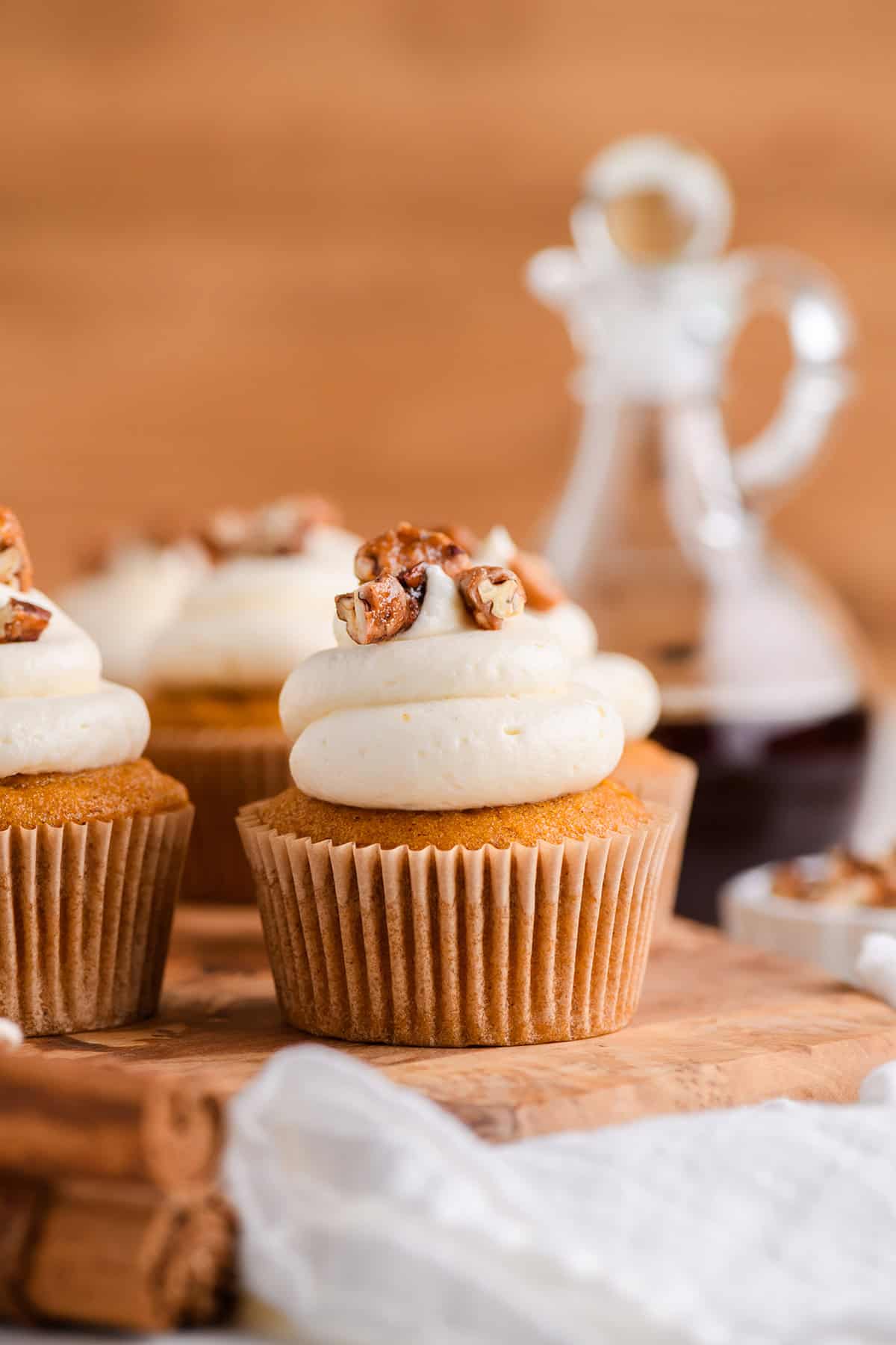 Close up shot of one cupcake on wood serving board with maple syrup jar in the background for Pumpkin Cupcakes with Maple Cream Cheese Frosting.