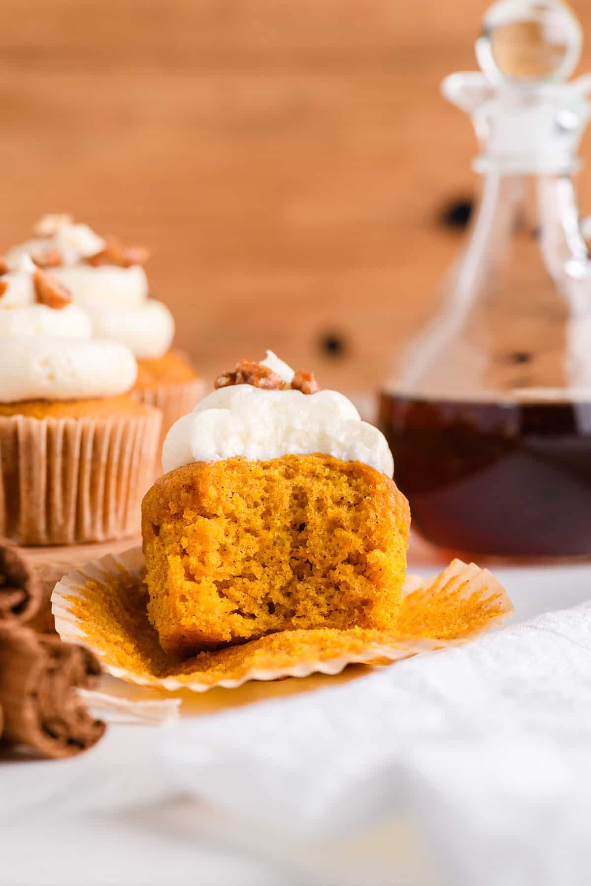 Close up of one Pumpkin Cupcakes with Maple Cream Cheese Frosting with bite and maple syrup jar in the background.