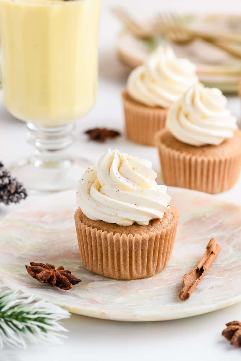 Wide open shot of one Eggnog Cupcake on a plate with a glass of eggnog and cupcakes in the background.