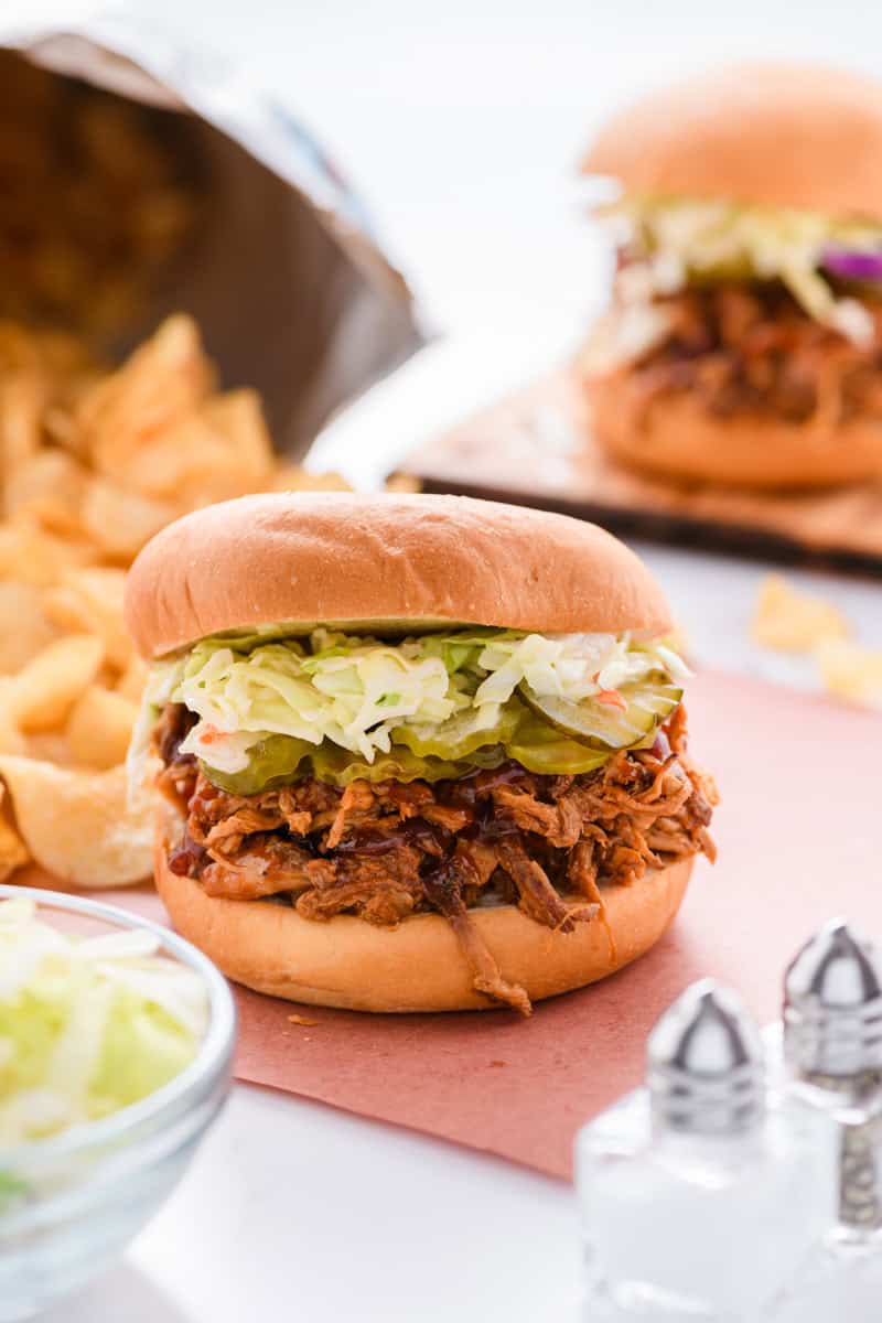 Close up shot of Slow Cooker Pulled Pork Sandwich with chips and second sandwich in the background.