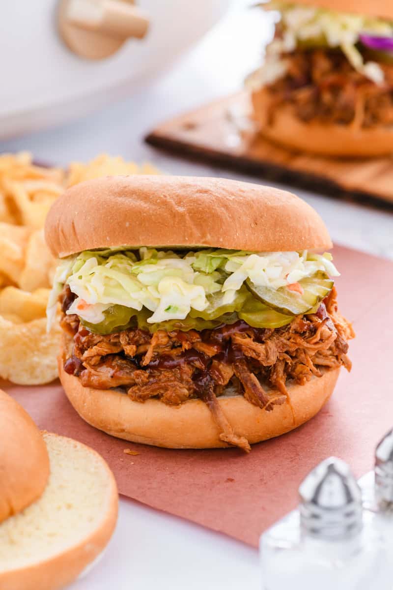 Close up shot of Pulled Pork Sandwich with chips and slow cooker in the background.
