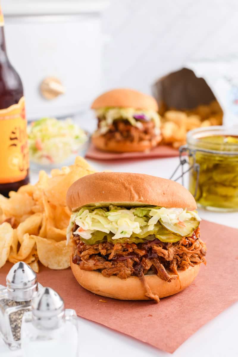 Wide open shot of Slow Cooker Pulled Pork Sandwiches with beer and slow cooker in the background.