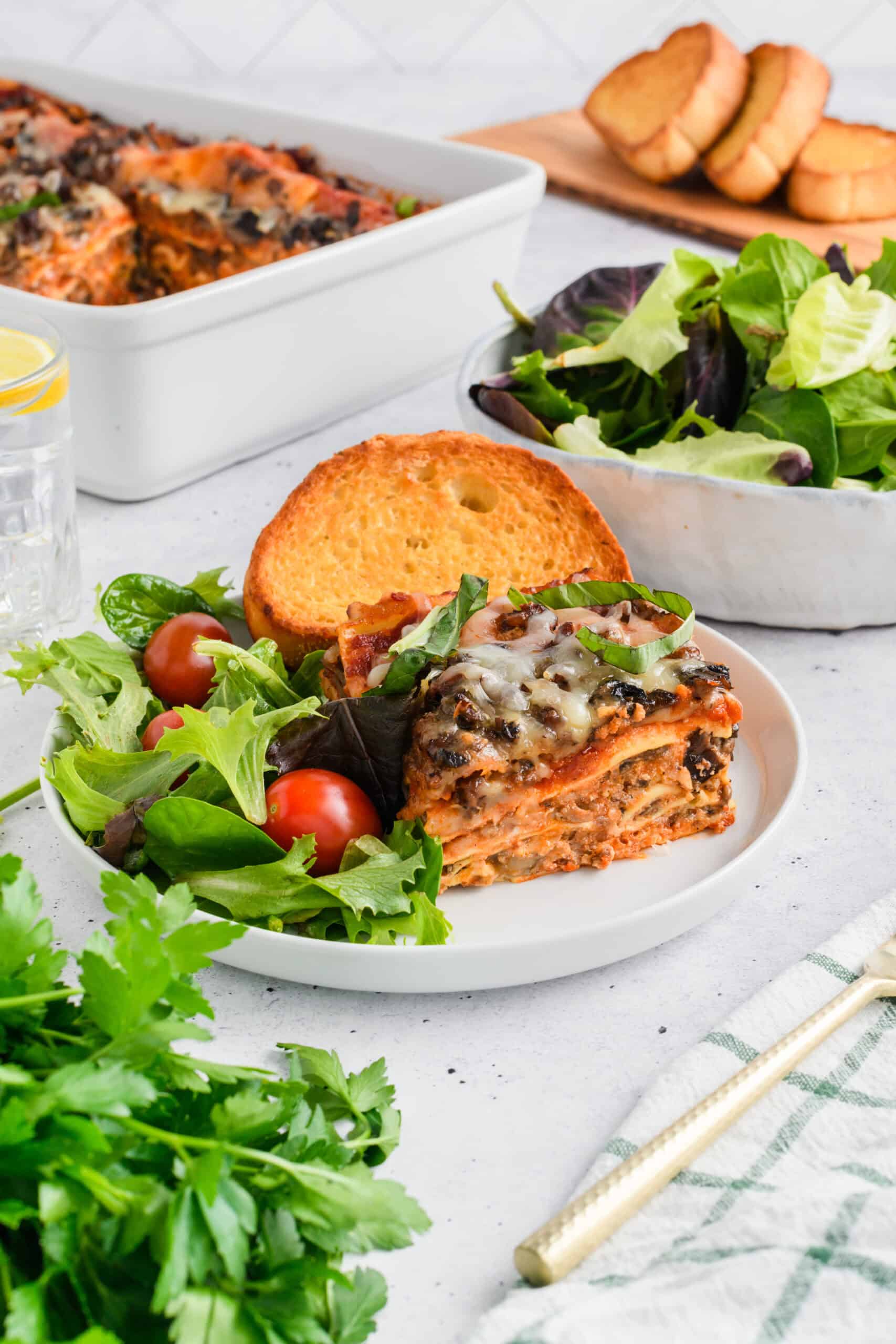 Wide open shot of served slice of Easy Sausage and Mushroom Lasagna with salad and garlic bread.