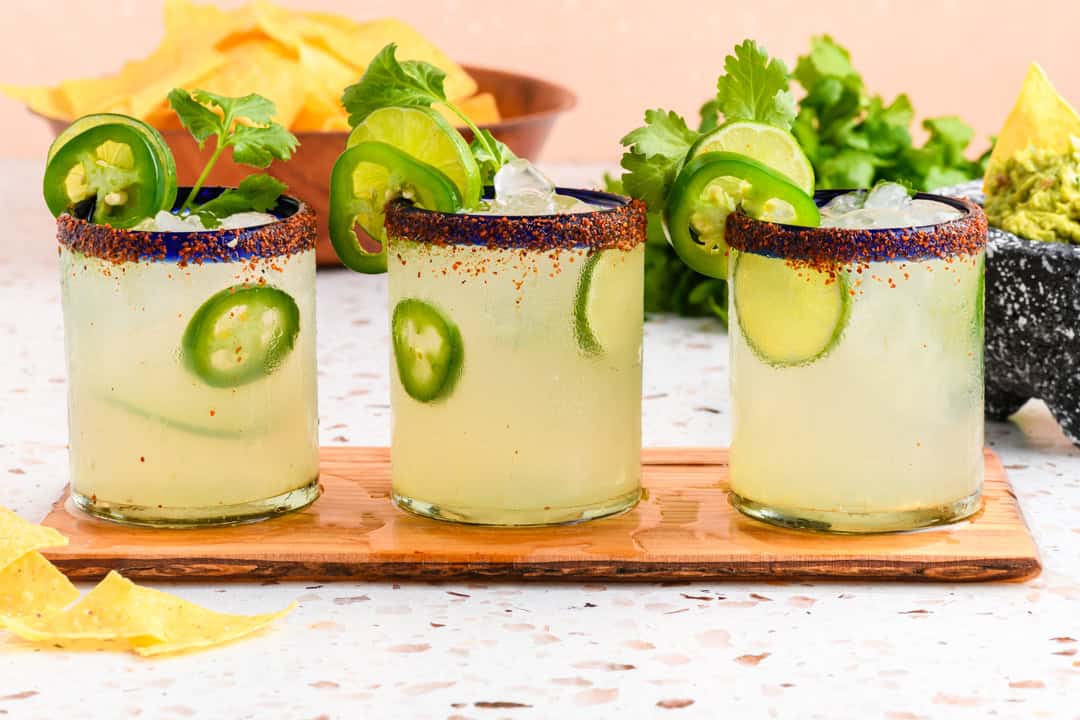Three Jalapeño Margaritas on wood serving tray with chips and guacamole nearby.