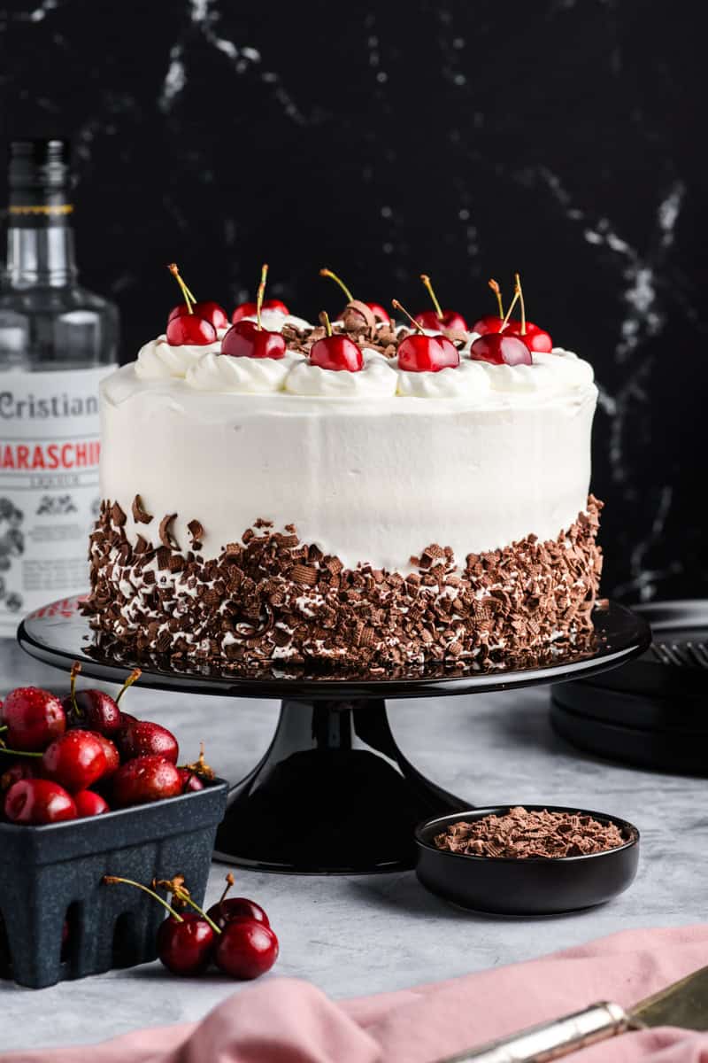 Black Forest Cake on black cake stand with crate of cherries and cherry liqueur.