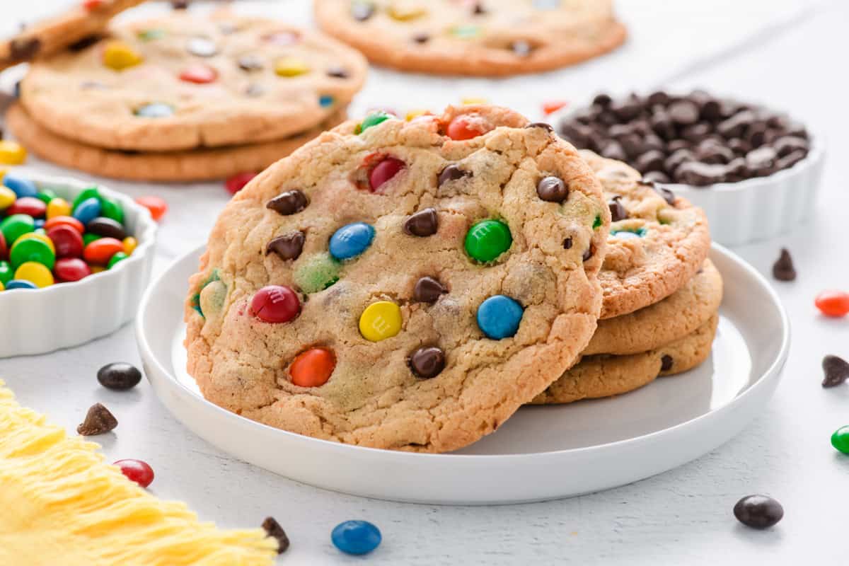 Angled shot of plate of stacked Best Big Fat Chewy Chocolate Chip M&M Cookies with hero cookie leaning up against the stack on a white plate.